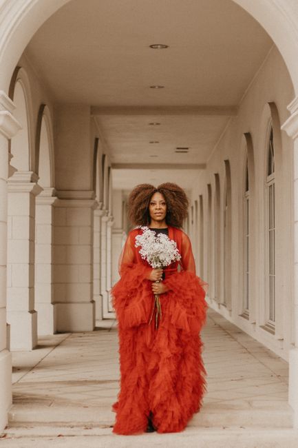 A stylish woman in a vibrant red gown holding a bouquet in an elegant corridor.