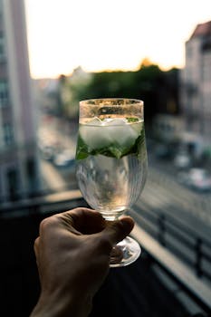 A hand holding a fresh cocktail with mint and ice on a balcony during sunset.