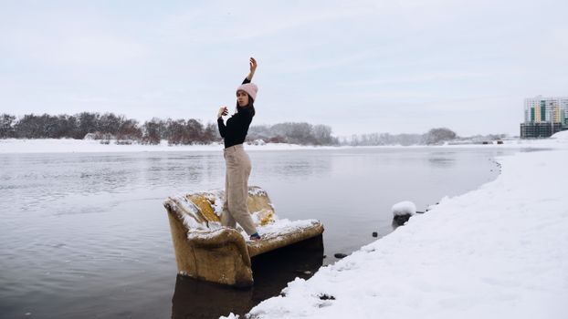 Woman standing on a snow-covered sofa by a river in winter, showcasing serenity and unique perspective.