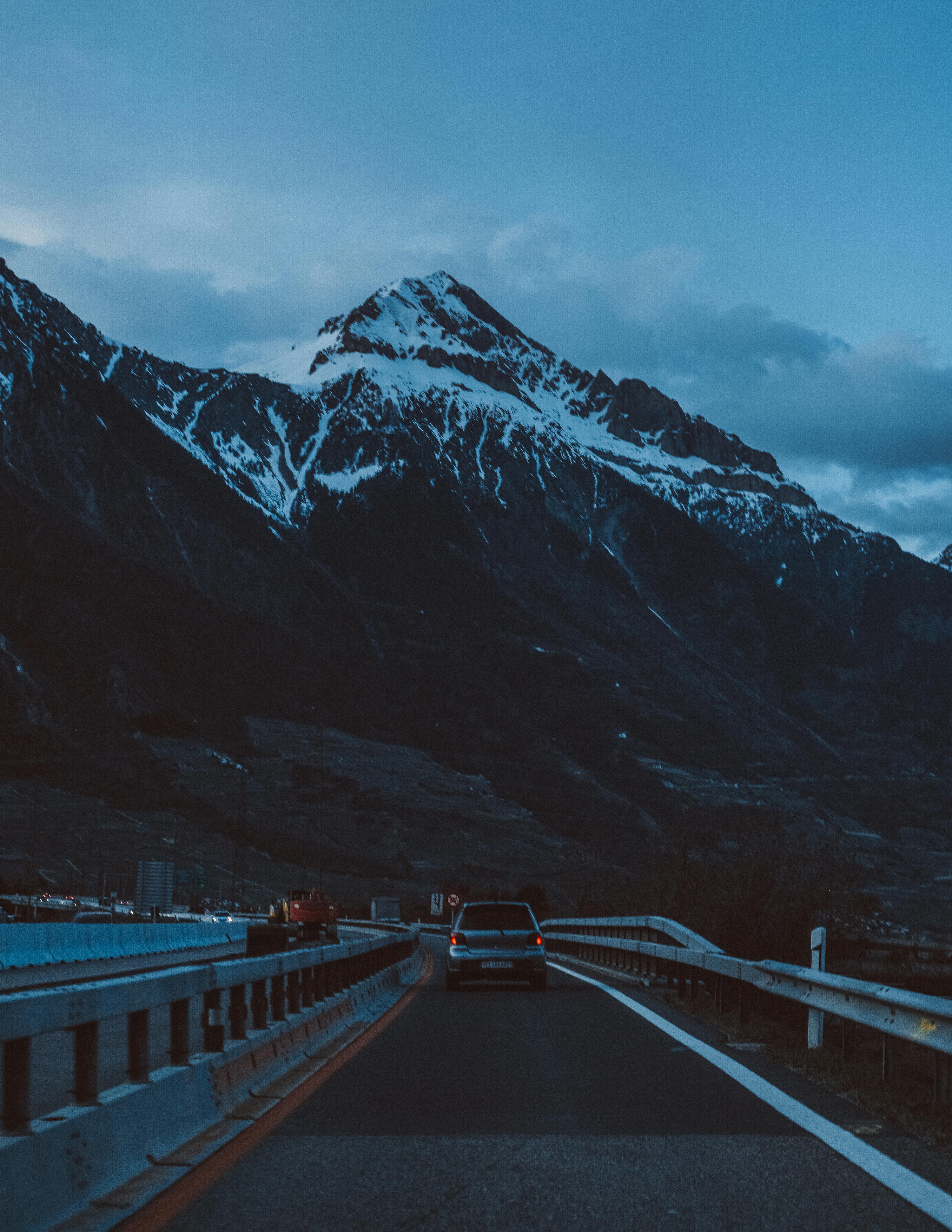 A car drives along a mountain road in Airolo, Switzerland at twilight, with snow-capped peaks in the background.