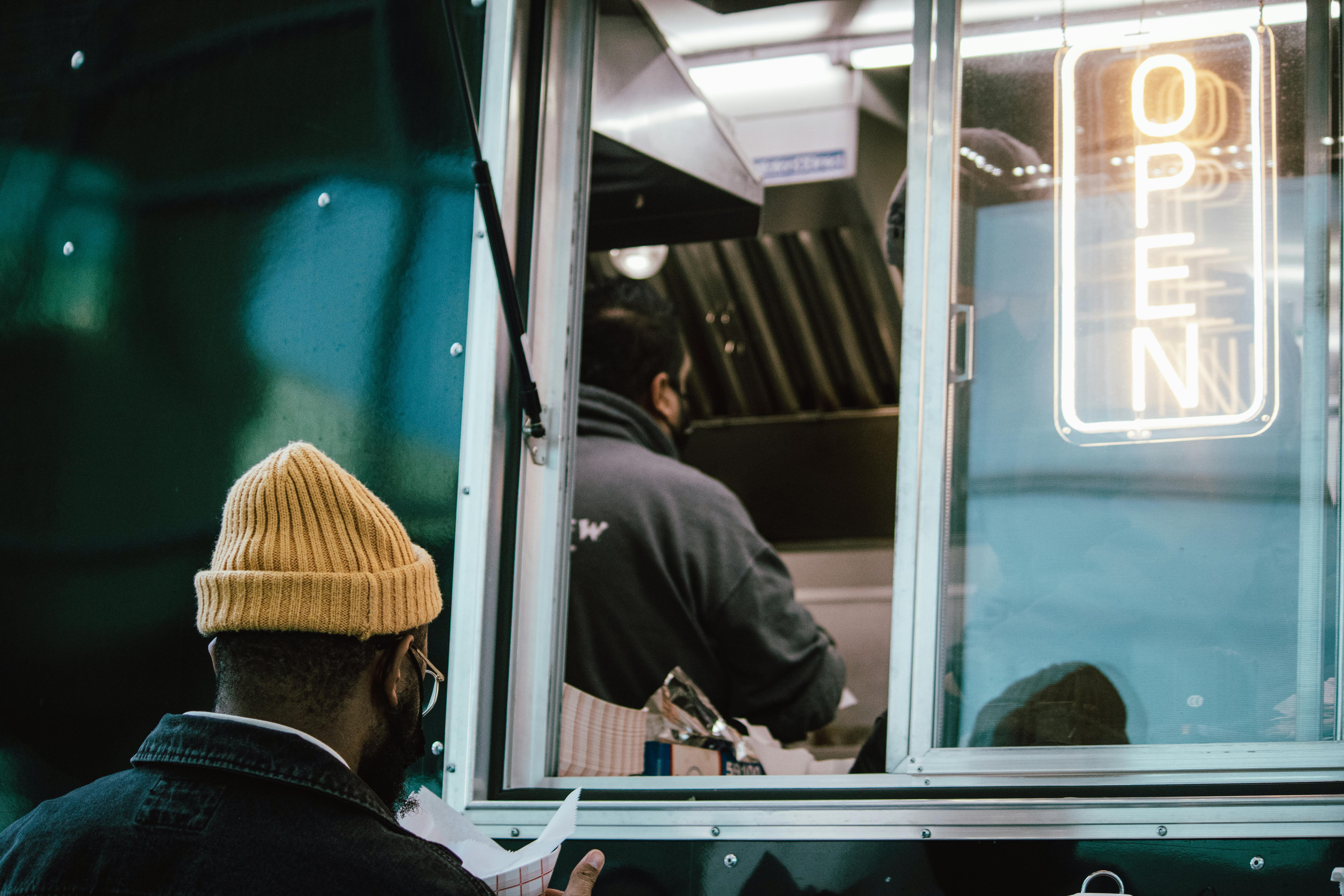 Man orders food from a food truck at night, neon 'open' sign glowing.