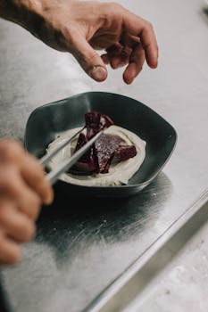 Close-up of a chef's hands garnishing a beetroot dish with sauce in a modern kitchen.