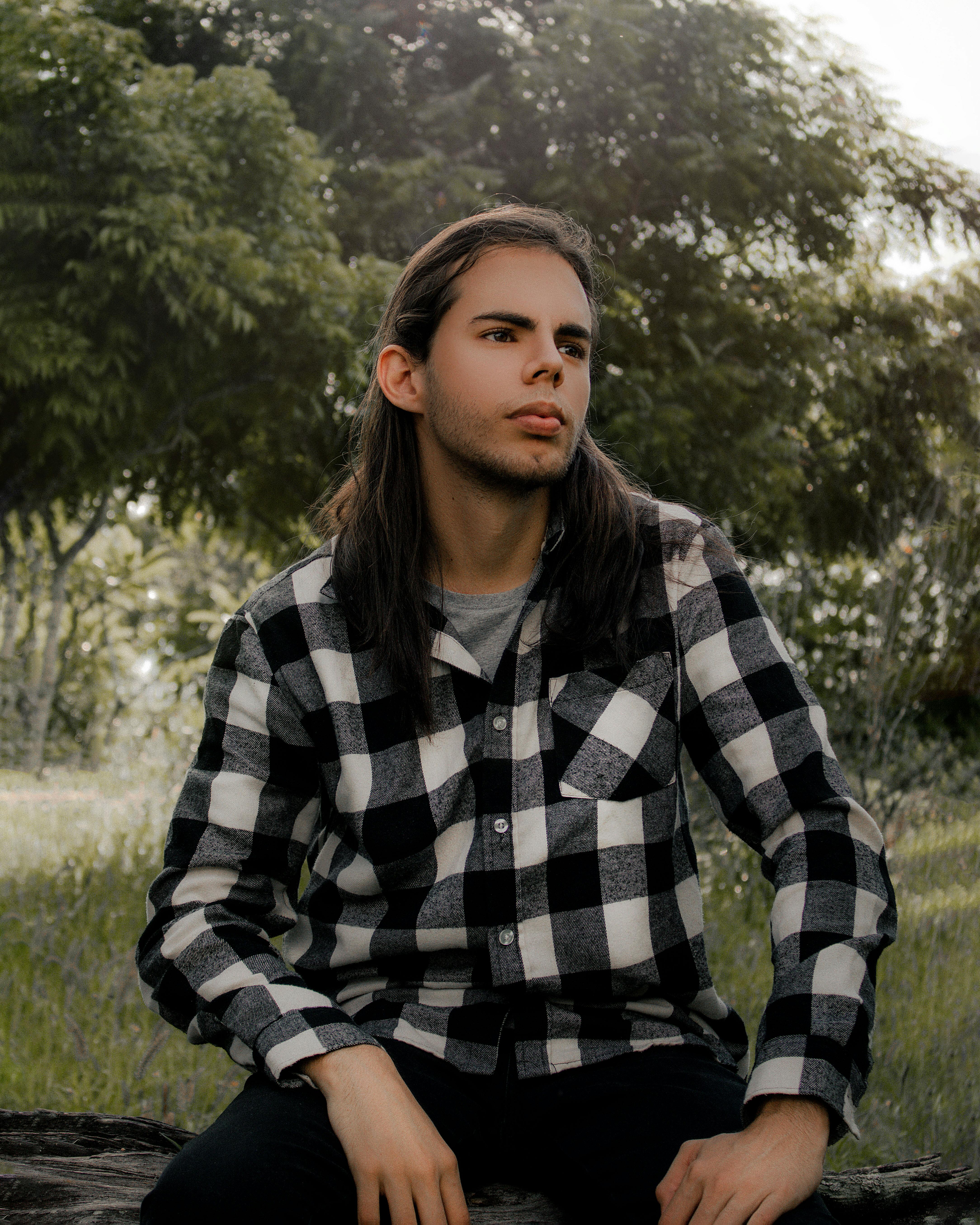 Portrait of a young man with long hair sitting in a park wearing a checked shirt.