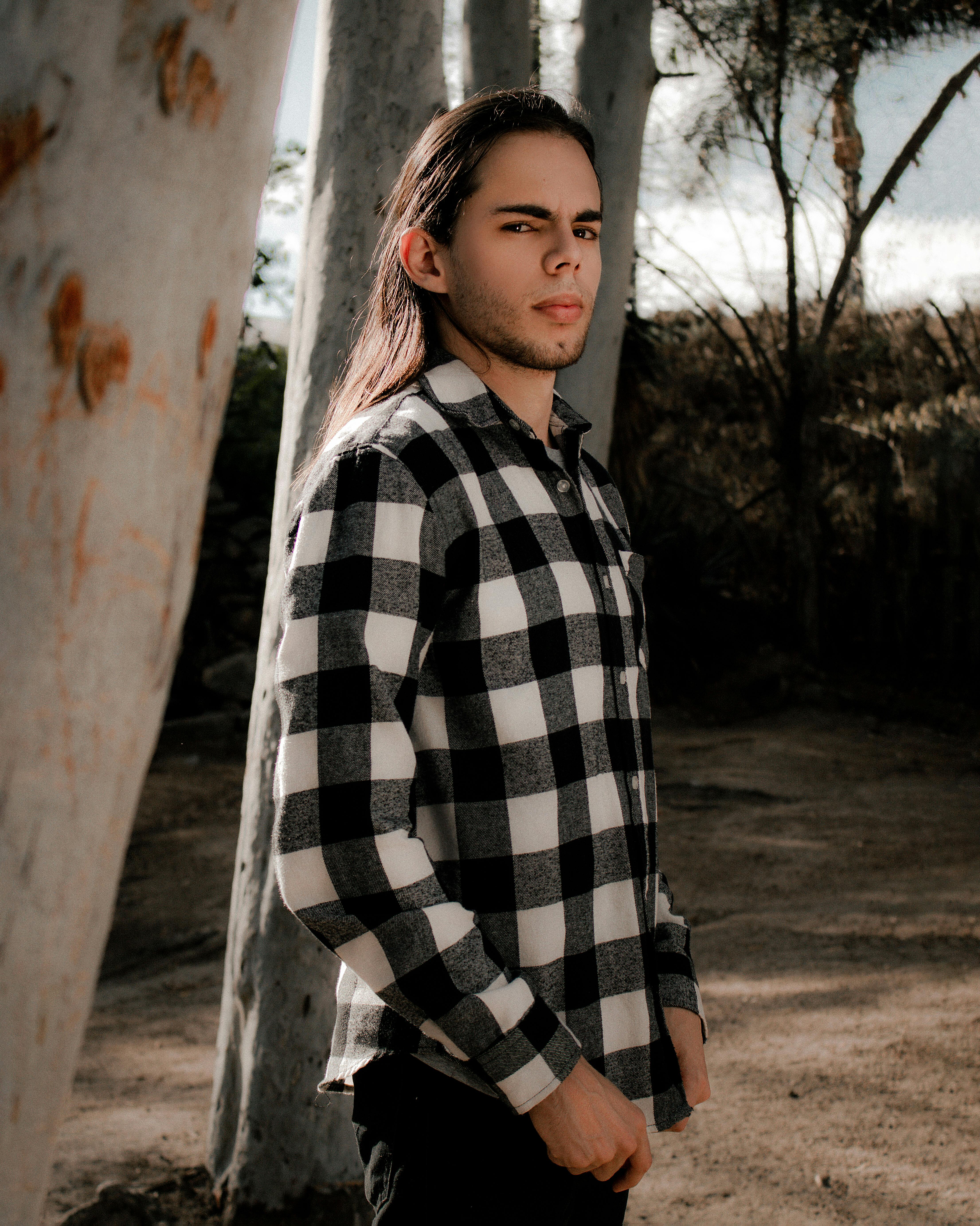Serene portrait of a young man in a checked shirt, standing amidst sunlit trees, exuding a calm and serious demeanor.