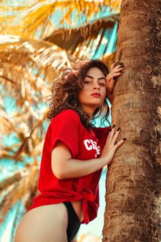 Woman in red shirt posing by palm tree with tropical background.