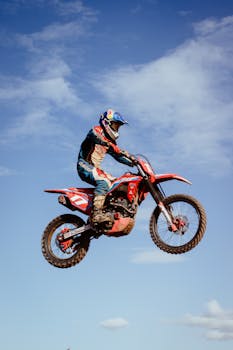 Rider performing a jump on a red motocross bike under a clear blue sky, showcasing dynamic motion.