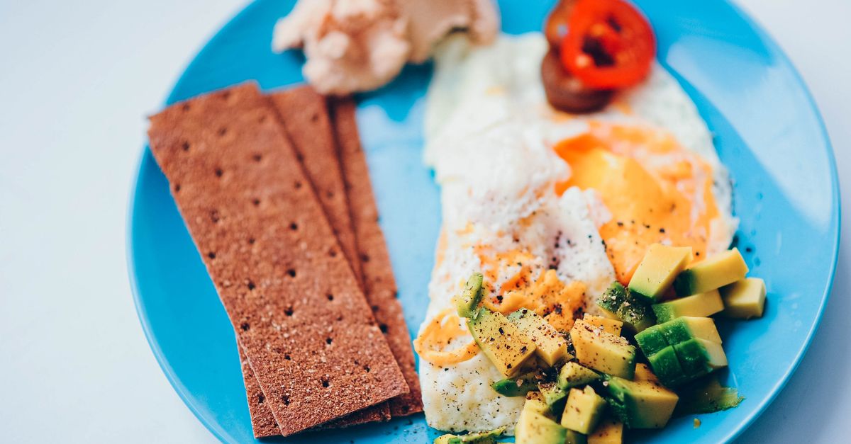 A close-up of a plate of waakye with shito, a fried egg, and sliced avocado, placed on a wooden table with a white church bulletin in the background