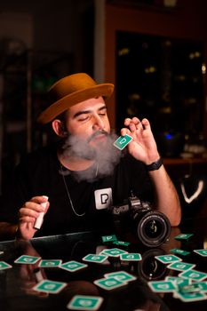 Man with brown hat vaping, holding cards in a dimly lit room, DSLR camera on table. 11121952