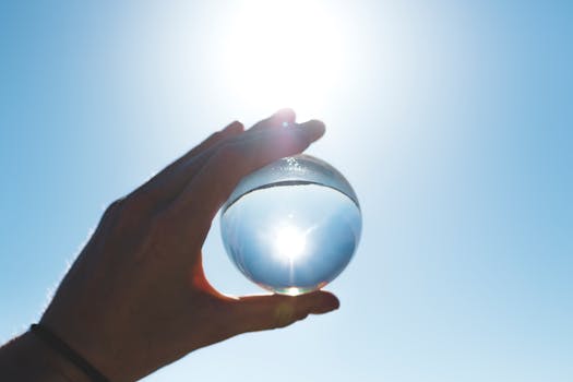 A crystal ball held by a hand reflects the bright sun in a clear blue sky.