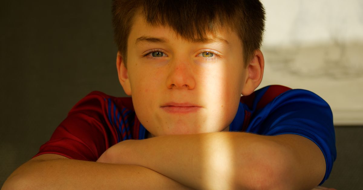 A serene portrait of a teenage boy resting his arms on a table indoors with natural light casting a warm glow.