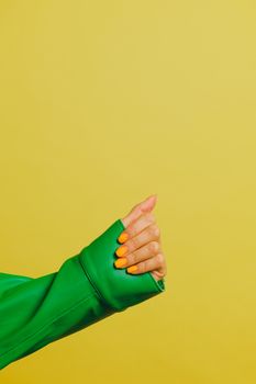 Close-up of a hand with orange nails and green jacket, vibrant yellow background.