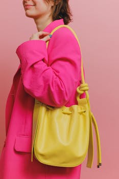 Fashionable woman in pink blazer holding yellow shoulder bag against pink background.