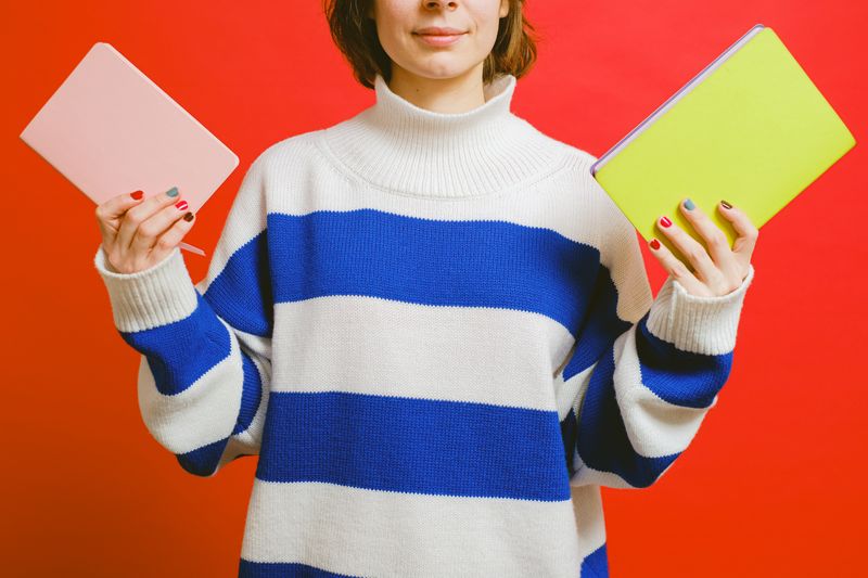 A woman holds pink and yellow notebooks while wearing a white and blue striped sweater against a red background.