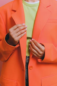 Close-up of hands adjusting a vibrant orange blazer over a green top, showcasing modern fashion.