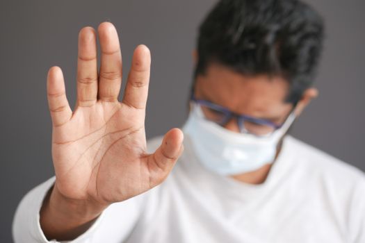 Close-up of a man wearing a face mask and eyeglasses, raising his hand in a stop gesture.