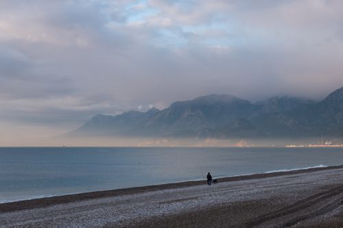 Free A solitary figure walking along a tranquil beach with distant misty mountains under a cloudy sky. Stock Photo