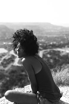 Black and white image of a woman with afro hair sitting on a mountain with a scenic view.