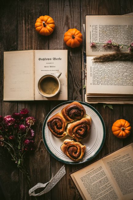 Cozy autumn setup with coffee, books, cinnamon rolls, and pumpkins on a wooden table.