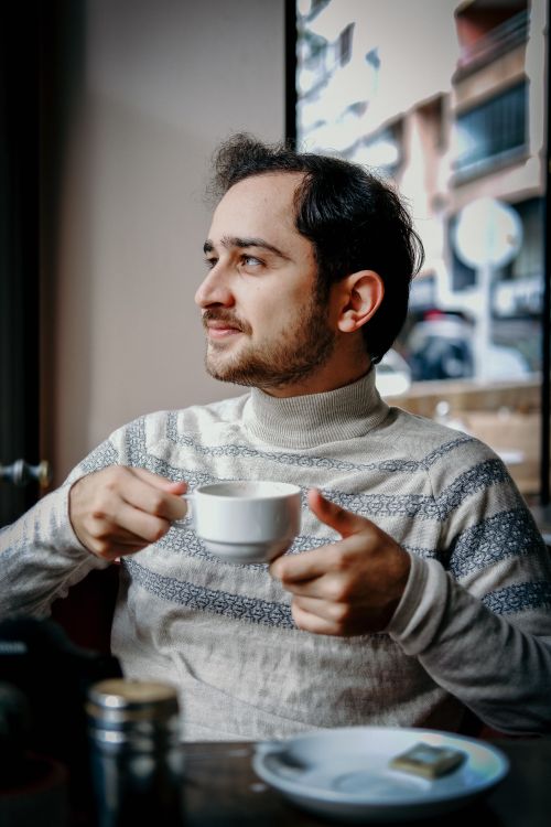 Free Young man with a mustache enjoying a coffee in a cozy indoor cafe, looking thoughtful. Stock Photo