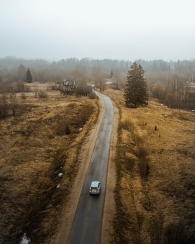 Aerial shot of a car driving on a rural road through a misty landscape with barren trees.
