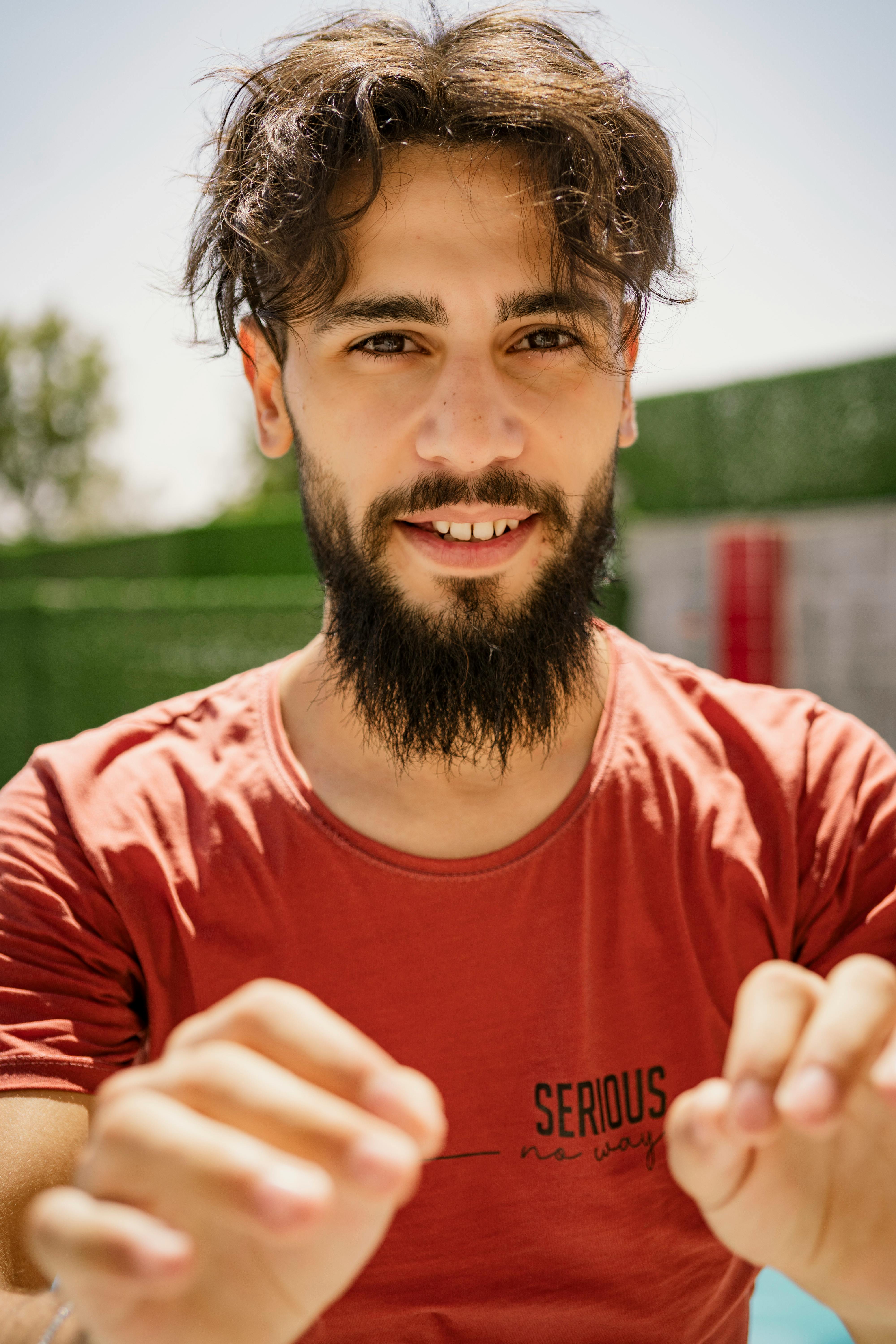 Smiling bearded man in a red shirt outdoors on a sunny day, showcasing facial expressions.