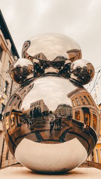 Modern reflective sculpture in urban setting with city architecture mirrored on surface.