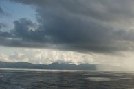 A moody seascape with dramatic clouds over a calm ocean and distant mountains.