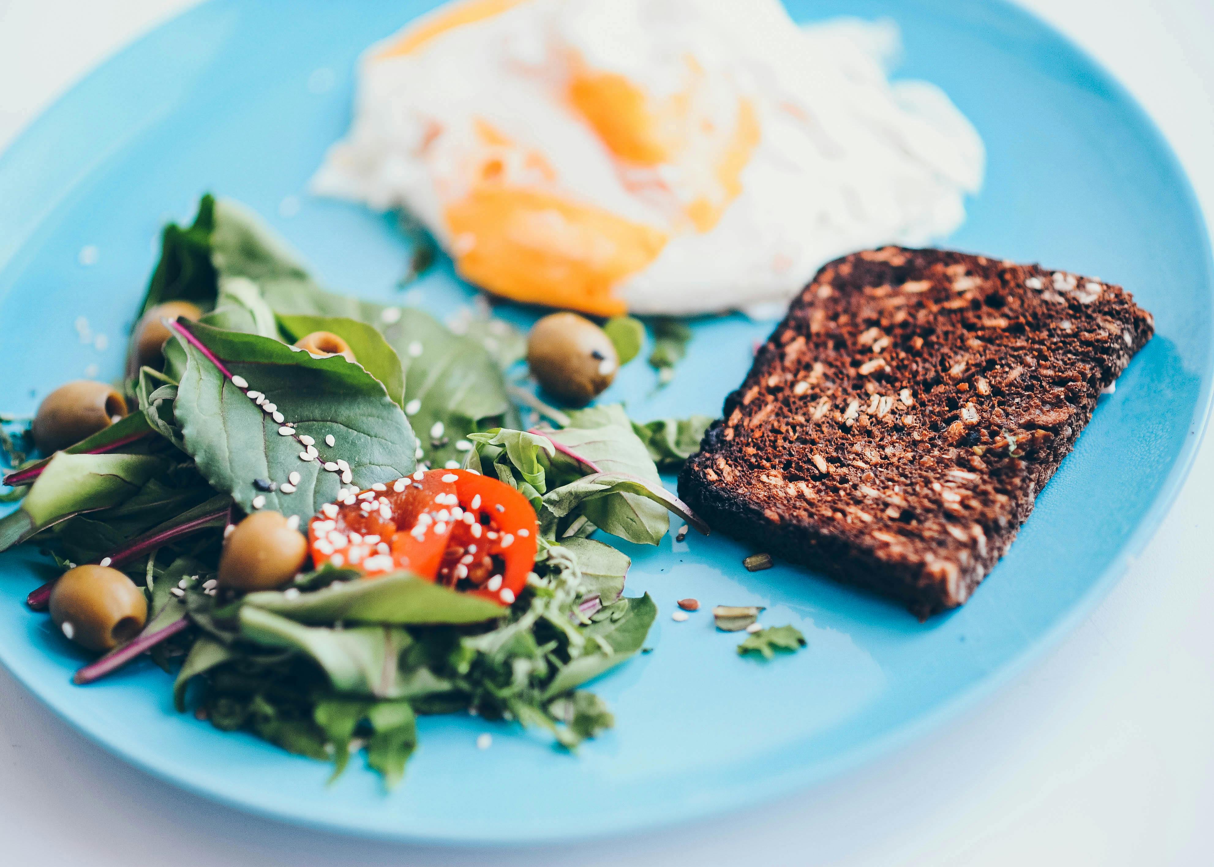 A nutritious breakfast plate featuring eggs, rye bread, salad, and olives on a vibrant blue plate.