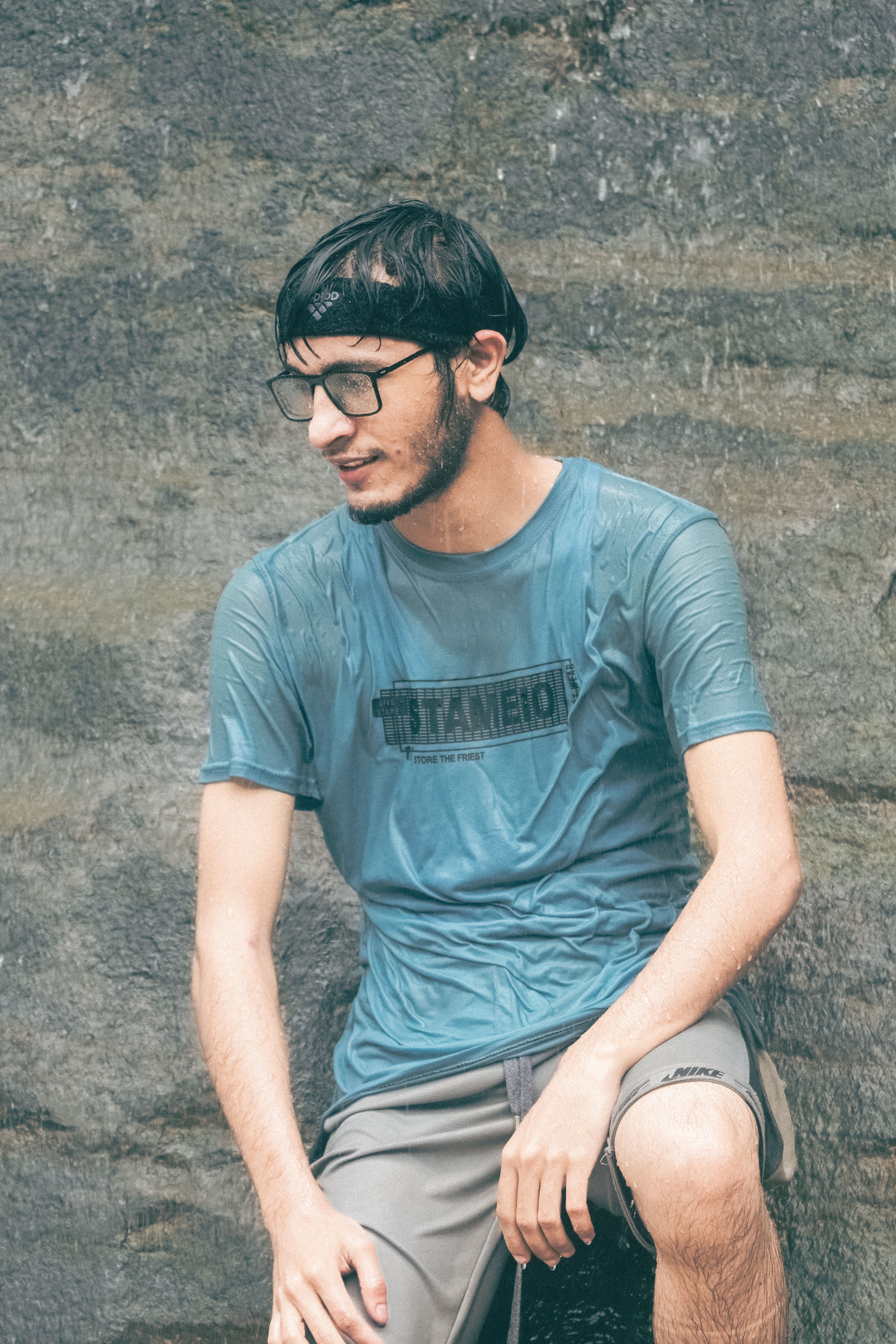 A young man in a wet blue shirt and glasses sits by a rock wall, enjoying the outdoors.
