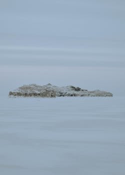 A remote snow-covered island emerges through the winter fog on a calm lake, creating a serene and isolated scene.
