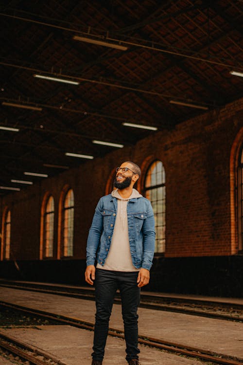 Free Smiling Man with Beard in Eyeglasses and Jean Jacket Standing on Train Tracks Stock Photo