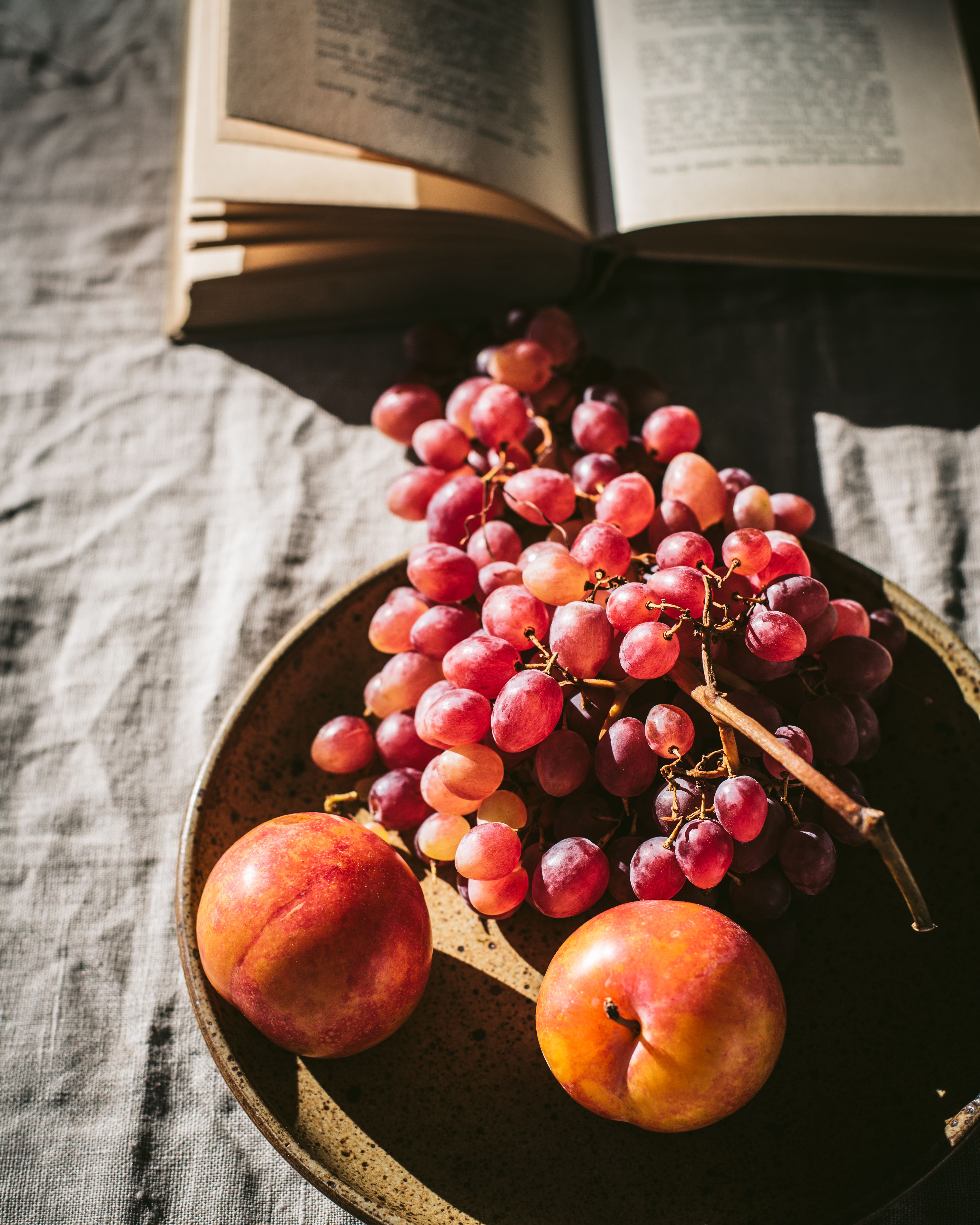 A rustic still life composition featuring fresh grapes, peaches, and an open book on a textured tablecloth.