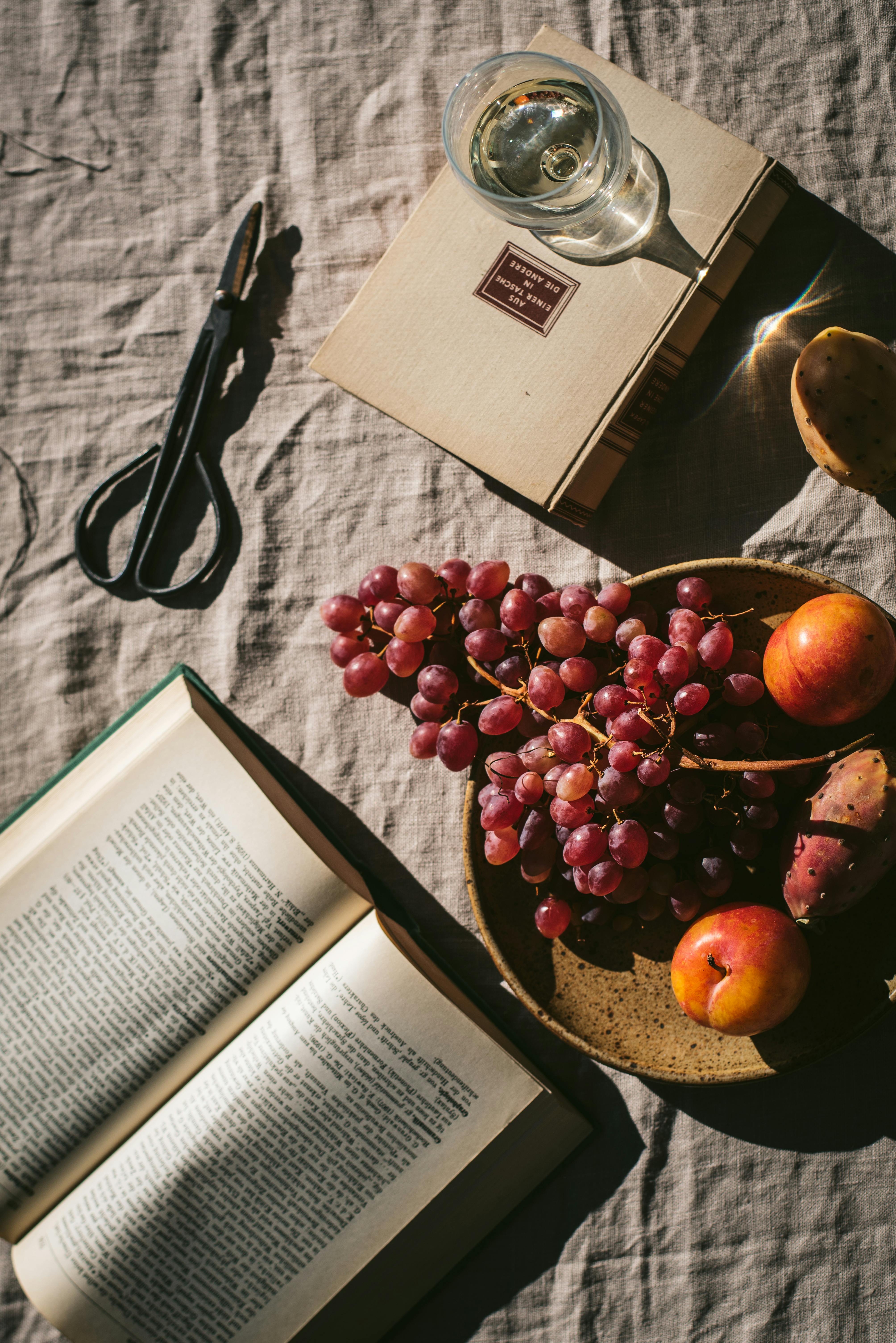 Stylish flat lay featuring wine, fruits, and books on a textured surface in warm sunlight.