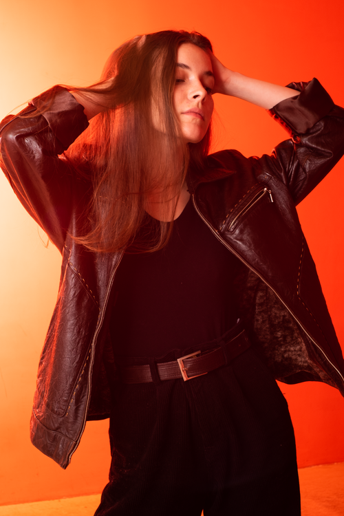 Free Woman in black leather jacket and pants posing against an orange backdrop with eyes closed. Stock Photo