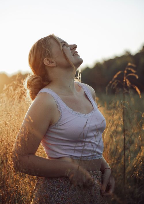 Free A woman in a white tank top smiles while standing in a sunlit field during sunset. Stock Photo