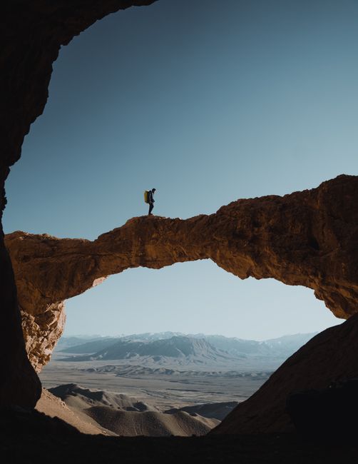 Silhouette of a lone traveler standing on a majestic natural rock bridge, with mountains in the background.
