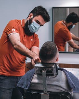 A barber in an orange shirt cutting a man's hair while wearing a face mask.