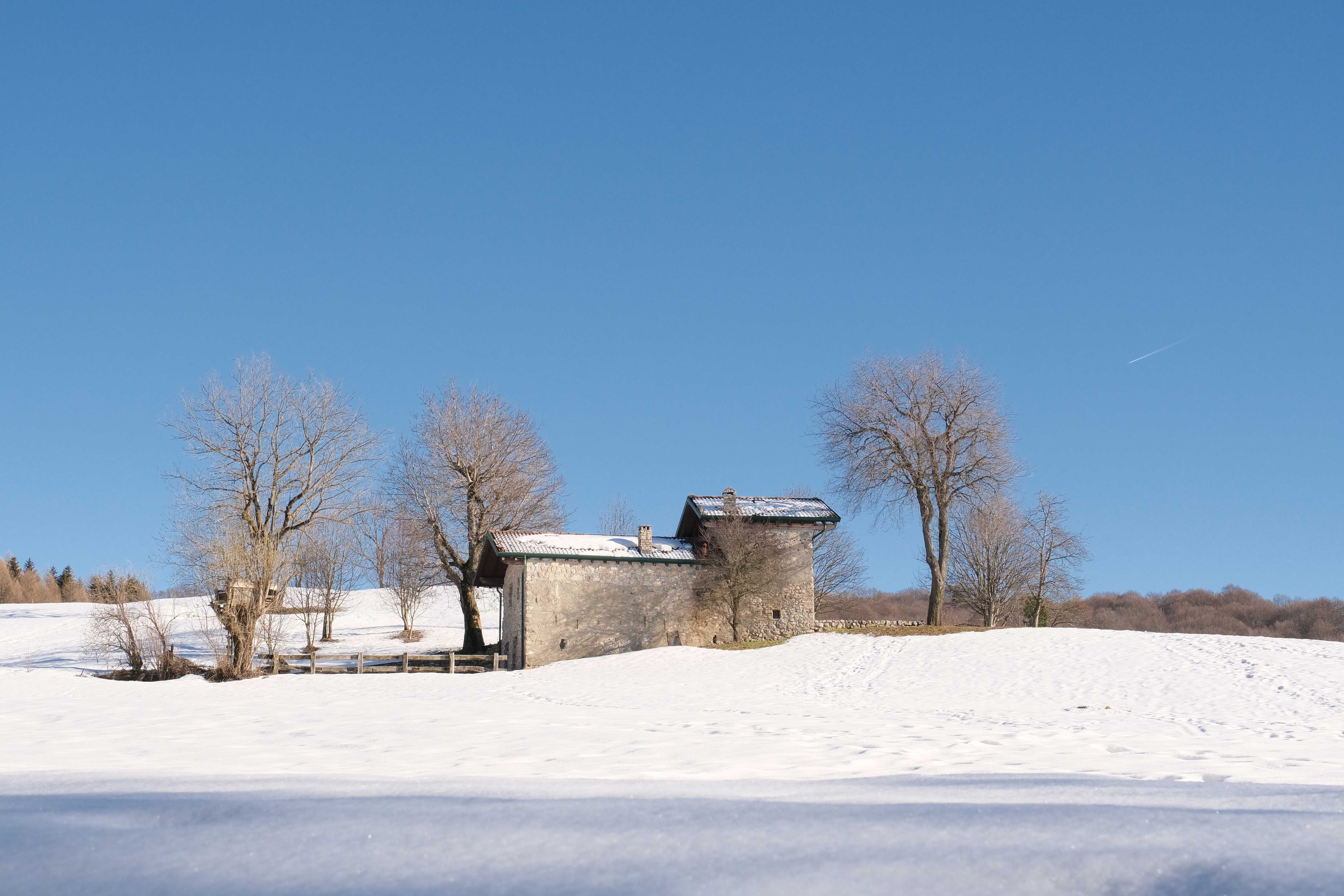 Stone house amid snow-covered fields and leafless trees under a clear blue sky.