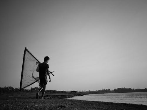 Grayscale Photo of Man Walking while Holding a Fish Net Near the Sea