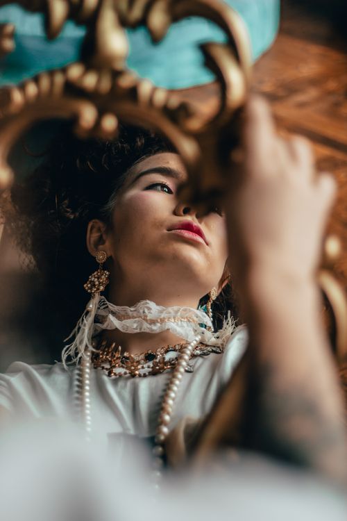 Free A woman with curly hair elegantly dressed in vintage style, observes her reflection in an ornate mirror. Stock Photo