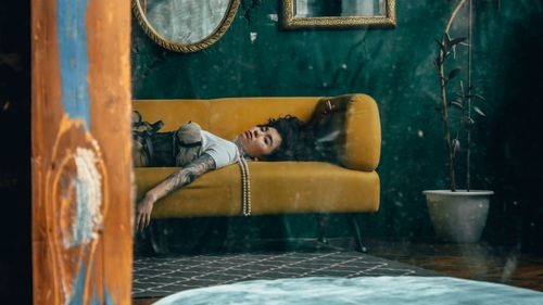 Free A woman with tattoos reclines on a vintage yellow sofa, reflecting modern classic style. Stock Photo