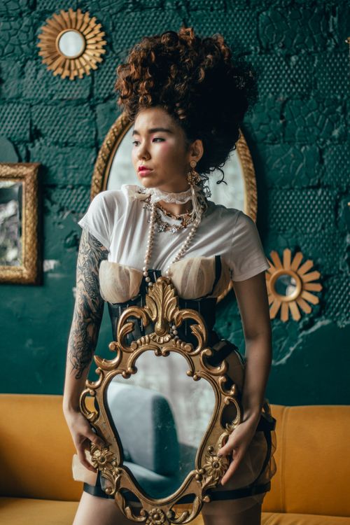 Free A stylish woman with intricate hair and tattoos holds an ornate mirror, standing against a decorative green wall. Stock Photo