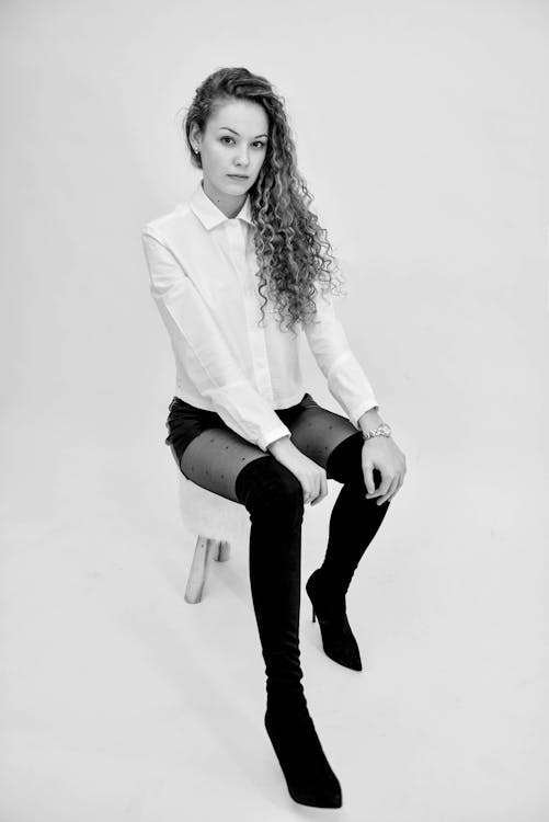 A Grayscale Photo of a Woman in White Long Sleeves Sitting while Wearing Black Boots