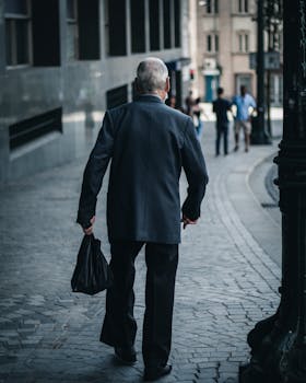 Mature man in suit walking on a city sidewalk, carrying a bag.