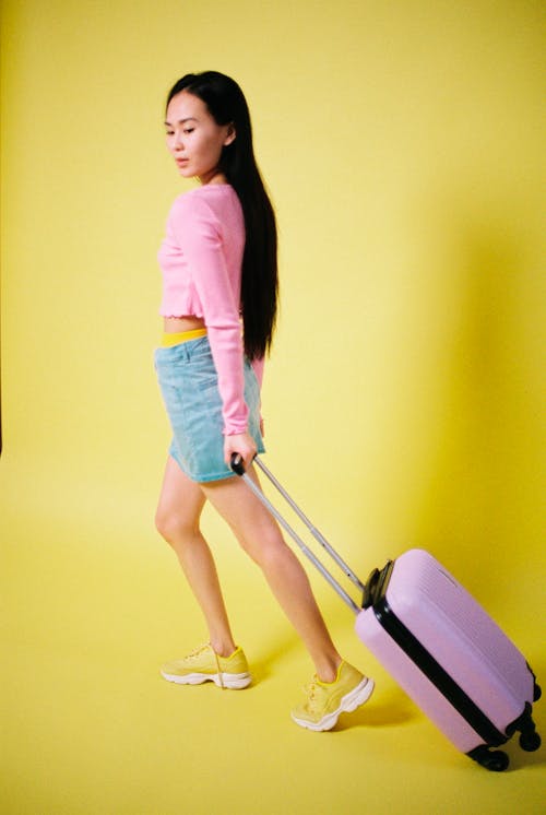 A Woman in Pink Long Sleeves and Denim Skirt Pulling a Luggage