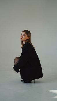 Studio portrait of a stylish woman crouching in a black jacket against a gray backdrop.