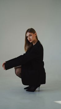 Well-dressed woman with long hair crouching in heels against a gray background, showcasing simplicity and style.