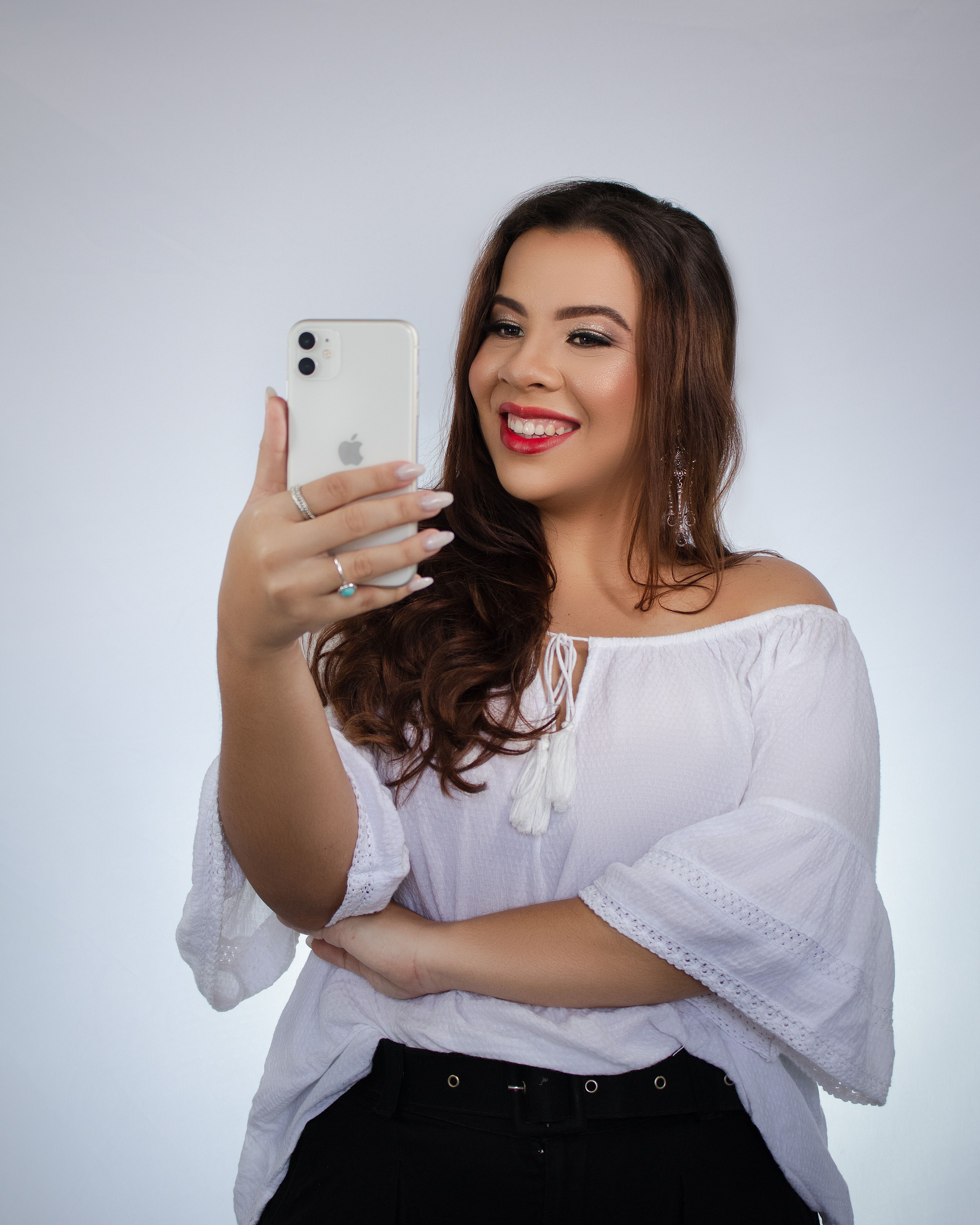 Cheerful woman in a white top smiling while taking a selfie with a smartphone against a white background.