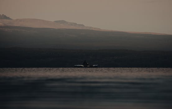 A lone motorboat floats tranquil on a calm lake under twilight skies, surrounded by distant hills.