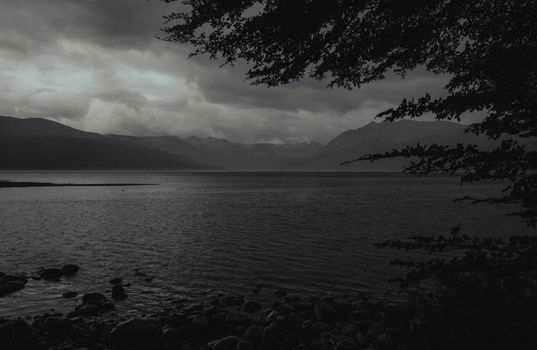 Serene monochrome lake with mountain backdrop and dramatic cloud cover.